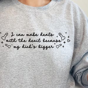 May include: A light gray sweatshirt with the text "I can make deals with the devil because my dick's bigger" in black script. The text is surrounded by small penis illustrations, hearts, and stars.