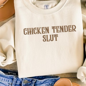 May include: Cream-colored sweatshirt with the phrase "CHICKEN TENDER SLUT" in brown lettering. The sweatshirt is laid on a surface with denim jeans, a brown hat, and sandals.
