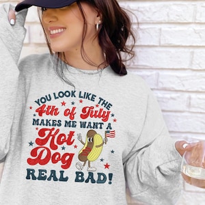 May include: A light gray sweatshirt with the text "YOU LOOK LIKE THE 4th of July MAKES ME WANT A Hot Dog REAL BAD!" in red, blue, and white. The design includes stars and a cartoon hot dog holding an American flag.