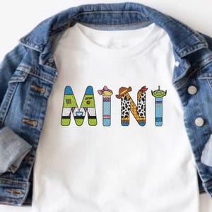 May include: White t-shirt with the word "MINI" spelled out in colorful letters. Each letter is decorated with a different character from the Toy Story movie franchise.