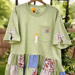 May include: A light green speckled t-shirt with a chest pocket and a Carhartt logo patch. The sleeves feature white fabric bows with floral patterns. The bottom hem has a ruffled patchwork skirt made of floral, blue polka dot, and beige gingham fabric.