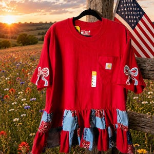May include: A red short-sleeve t-shirt with a chest pocket featuring a Carhartt logo patch. The sleeves are accented with white and blue bow appliqués. The hem features a ruffled patchwork trim in red, white, and blue patterns.