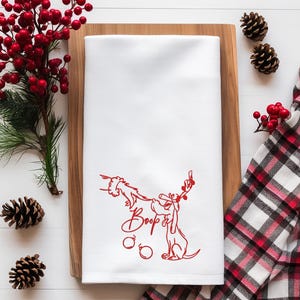 May include: White kitchen towel with a red line drawing of a dog wearing antlers and holding a Christmas ornament. The word "Boop" is written in red script below the dog. The towel is on a wooden cutting board.