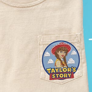 May include: A beige t-shirt with a chest pocket featuring an embroidered circular patch. The patch depicts a person with braided hair wearing a red cowgirl hat and yellow shirt, set against a blue sky with white clouds and the text Taylor's Story.