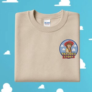 May include: A beige crewneck sweatshirt featuring an embroidered circular patch on the left chest. The patch depicts a cartoon character wearing a red cowgirl hat, set against a blue sky with white clouds, and the text "TAYLOR'S STORY" in yellow.