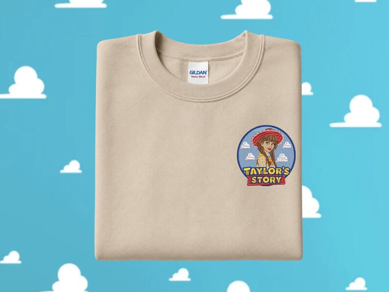 May include: A beige crewneck sweatshirt featuring an embroidered circular patch on the left chest. The patch depicts a cartoon character wearing a red cowgirl hat, set against a blue sky with white clouds, and the text "TAYLOR'S STORY" in yellow.
