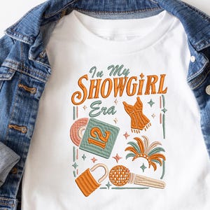 Könnte beinhalten: Weißes T-Shirt mit dem Text "In My Showgirl Era" in Orange und Grün. Das Design umfasst ein Korsett, eine Schallplatte, ein Schloss, ein Mikrofon und eine Federboa. Das Shirt wird auf einer Jeansjacke präsentiert.