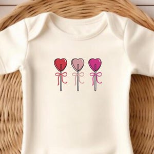 May include: White baby onesie with a graphic of three heart-shaped lollipops on sticks. The lollipops are red, pink, and light pink.