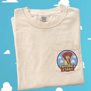 May include: A cream-colored short-sleeve t-shirt with a chest pocket. The pocket features a circular graphic of a person wearing a red cowboy hat against a blue sky with white clouds. The text "TAYLOR'S STORY" is printed in yellow and red letters.