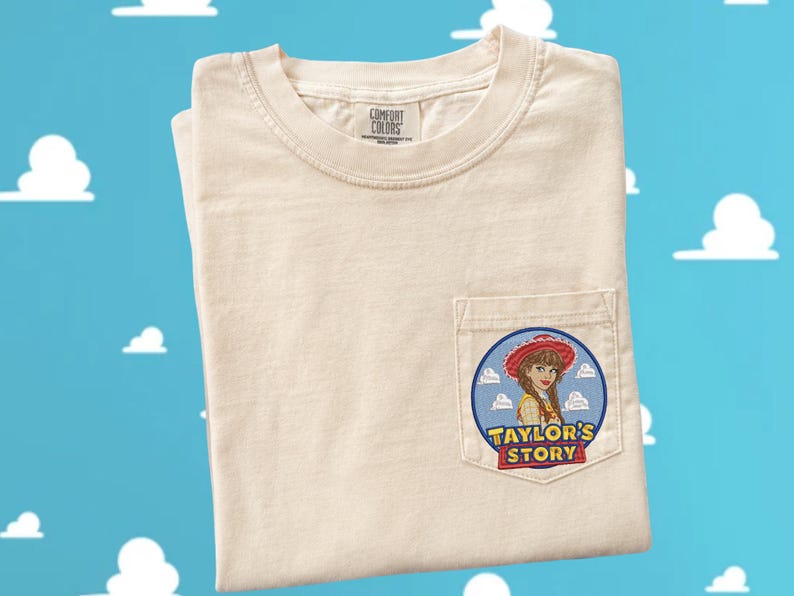 May include: A cream-colored short-sleeve t-shirt with a chest pocket. The pocket features a circular graphic of a person wearing a red cowboy hat against a blue sky with white clouds. The text "TAYLOR'S STORY" is printed in yellow and red letters.