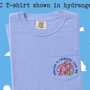 May include: A light purple short-sleeve t-shirt with a chest pocket. The pocket features a red and blue graphic of two figures with the text "YOU'RE A COWGIRL LIKE ME" and "TS". The inner neck label reads "COMFORT COLORS". Text above reads "CC T-shirt shown in hydrangea".