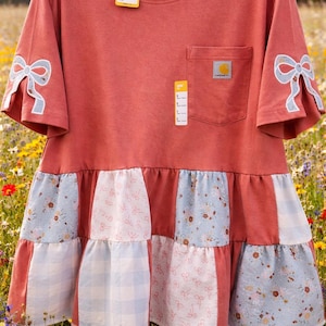 May include: A coral-colored t-shirt dress with a pocket and a Carhartt logo patch. The sleeves feature light blue bow accents. The dress has a patchwork skirt with floral and gingham patterns.