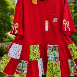 May include: A red short-sleeve t-shirt with a chest pocket and a Carhartt logo patch. The sleeves feature green and white bow graphics. The bottom hem has a tiered, ruffled skirt made of red fabric and patterned fabric with Grinch illustrations.
