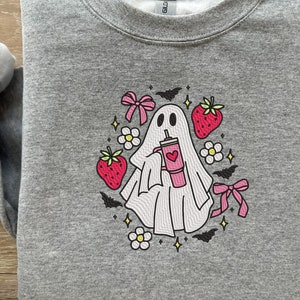 Puede incluir: Una sudadera gris con un fantasma blanco que sostiene una bebida rosa con un corazón. El fantasma está rodeado de lazos rosas, flores blancas y murciélagos negros.