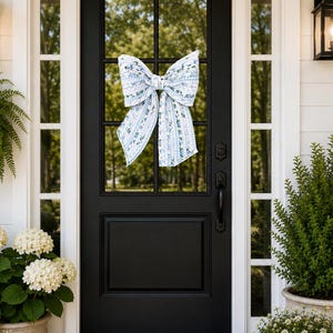 May include: A large decorative bow featuring a blue and white floral pattern with vertical stripes, attached to the glass pane of a black front door. The bow has two wide loops and two long, flowing tails, displaying a classic fabric texture.