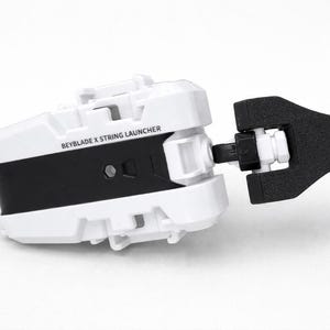 Può includere: Un lanciatore a corda Beyblade X bianco e nero. Il lanciatore ha un corpo rettangolare con la scritta "Beyblade X String Launcher" stampata sul lato. Il lanciatore ha un'impugnatura nera.