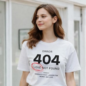 May include: White t-shirt with the text "ERROR 404 LOVE NOT FOUND" in black, with a red circle around the text. The shirt also has the text "SORRY! SORRENTLY BUSY LOOKING FOR MONEY, NOT LOVE" in black. The model is wearing khaki trousers.