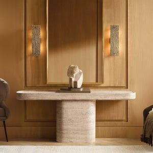 Puede incluir: Una consola de piedra beige con una parte superior redondeada y una base cilíndrica. Una pequeña escultura abstracta se encuentra encima. Dos apliques de pared rectangulares con superficies texturizadas flanquean un gran espejo enmarcado.