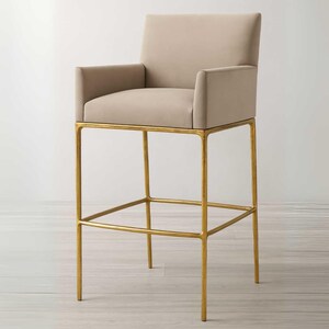 Può includere: Sgabello da bar imbottito beige con braccioli. La seduta e lo schienale sono di colore neutro e il telaio è in metallo color oro. Lo sgabello ha un poggiapiedi ed è progettato per una cucina o un bar.