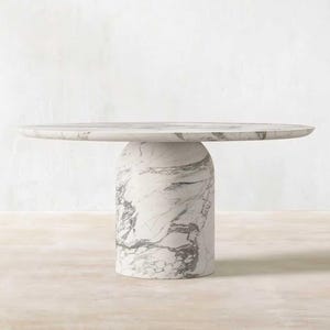 May include: A round marble table with a cylindrical base. The table is white with gray veining, and the top is approximately 48 inches in diameter. The base is a solid, rounded column. The table is suitable for a dining room or living room.