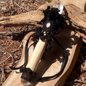Puede incluir: Una varita de madera hecha a mano con una punta de cristal de cuarzo transparente. La varita presenta detalles de ganchillo negro, una cuenta iridiscente central y cintas negras. La varita se exhibe sobre una superficie de madera, mostrando su estética natural.