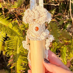 Puede incluir: Una varita de cristal de cuarzo transparente con un adorno de ganchillo color crema y un mango de madera. La varita presenta una pequeña piedra redonda de color marrón anaranjado en el centro del diseño de ganchillo. El fondo es follaje verde borroso.