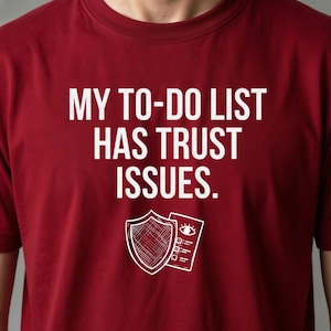 Könnte beinhalten: Ein kastanienbraunes Kurzarm-T-Shirt mit dem weißen Text "MY TO-DO LIST HAS TRUST ISSUES." Unter dem Text befindet sich eine weiße Strichgrafik, die einen Schild neben einer Checkliste mit einem Auge-Symbol am oberen Rand darstellt.