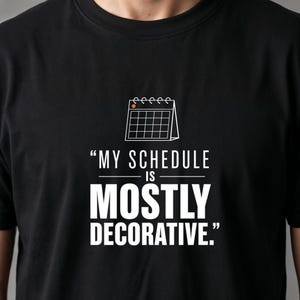 May include: A black crew neck t-shirt featuring a white graphic of a desk calendar with a small orange dot on the top left corner. Below the calendar, the text reads: "MY SCHEDULE IS MOSTLY DECORATIVE." in a bold, white, sans-serif font.