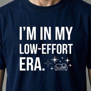 Può includere: Una t-shirt blu navy con il testo bianco "I'M IN MY LOW-EFFORT ERA." accompagnato da una piccola grafica di un divano con una coperta e diverse icone di stelle a quattro punte disposte attorno all'illustrazione del mobile.