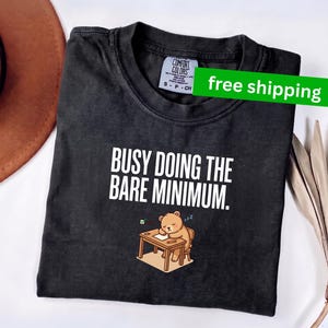 May include: A charcoal gray short-sleeve t-shirt with a crew neckline. The shirt features the white text "BUSY DOING THE BARE MINIMUM." above a graphic of a light brown bear sleeping at a wooden desk with a green checkmark and blue "zzz" symbols.
