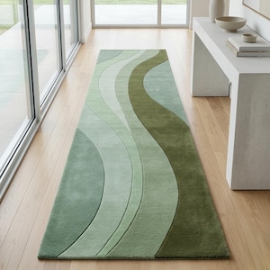 May include: A long, rectangular rug with a flowing, wave-like design in shades of green. The rug features a gradient of colours, from light green to a darker olive green. The rug is placed on a light-coloured wooden floor.