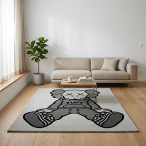 K&ouml;nnte beinhalten: Rechteckiger grauer Teppich mit einem gro&szlig;en Cartoon-Charakter-Design in Grau- und Schwarzt&ouml;nen. Die Figur tr&auml;gt einen Kapuzenpullover mit dem Wort "TUFT" und Turnschuhen. Der Teppich befindet sich in einem Wohnzimmer.