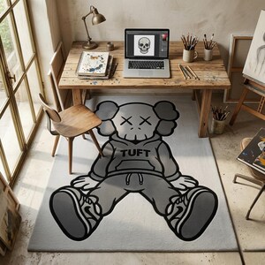 K&ouml;nnte beinhalten: Grau-wei&szlig;er Teppich mit einem Cartoon-Charakter mit durchgestrichenen Augen und dem Wort "TUFT" auf dem Kapuzenpullover. Der Teppich liegt auf einem hellen Boden, mit einem Holztisch und Laptop im Hintergrund.