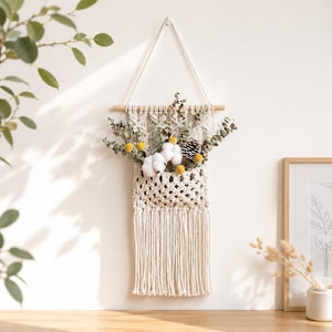 May include: A cream-colored macrame wall hanging featuring a woven pocket suspended from a wooden dowel with a white cord. The pocket contains dried eucalyptus branches, white cotton bolls, yellow billy balls, and a small brown pinecone.