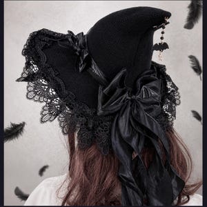 May include: A black knit witch hat featuring a wide brim with intricate black lace trim. The hat is adorned with a large black satin ribbon bow and a dangling charm featuring a small black bat and beads hanging from the pointed crown.