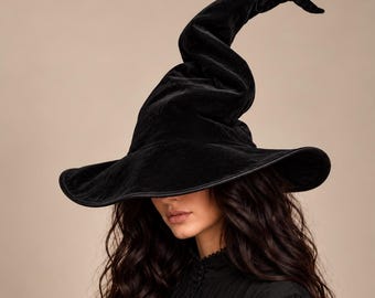 Puede incluir: Un sombrero de bruja de terciopelo negro con una copa alta, puntiaguda y ligeramente curvada. El sombrero presenta un ala ancha y flexible con un borde acabado. El material tiene una textura suave y reflectante, con una punta caída distintiva.