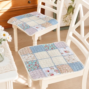 May include: Two square chair cushions featuring a quilted patchwork design with various floral and gingham patterns in shades of blue, pink, white, and green. The cushions are placed on white wooden chairs with a lattice backrest design.