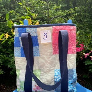 May include: A quilted tote bag featuring a patchwork design of blue, pink, green, and white fabric squares. The bag has two dark blue canvas straps and a white tag pinned to the front with the number 3 written in blue ink on the surface.