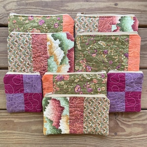May include: A collection of seven rectangular quilted zipper pouches in various sizes, featuring patchwork designs with floral, scalloped, and solid fabric patterns in shades of green, orange, purple, and pink with visible stitching.