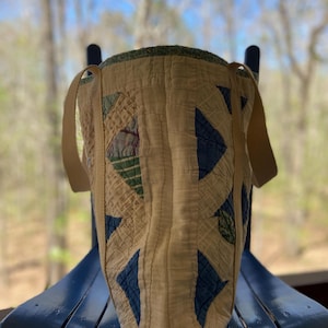 May include: A handmade tote bag with a quilted design. The bag features a cream-colored background with blue and green geometric patterns. It has two beige straps and a green trim around the top. The bag is sitting on a blue chair.