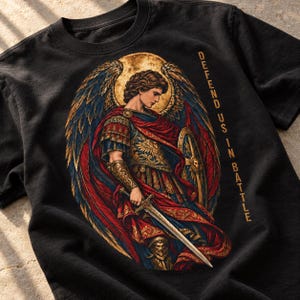 May include: A black t-shirt featuring a graphic of an angel in armour with large wings, holding a sword and shield. The angel wears a red cloak and gold armour. The text "DEFEND US IN BATTLE" is printed vertically in gold letters on the right side.