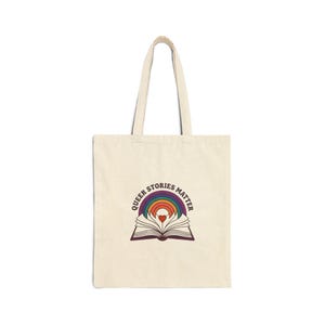 May include: Cream-colored tote bag with a rainbow and open book graphic. The text "QUEER STORIES MATTER" is arched above the rainbow. The bag has two long handles for carrying over the shoulder.