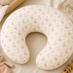 Puede incluir: Una almohada de lactancia de color crema con forma de U, cubierta con un patrón repetido de pequeños conejitos de dibujos animados en rosa y beige. También se ven un juguete de conejo de ganchillo y una sonaja de madera, lo que sugiere un tema de bebé o guardería.