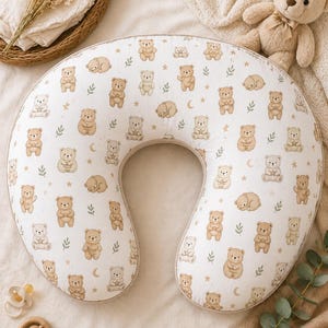 May include: A white nursing pillow with a pattern of tan teddy bears, stars, and crescent moons. The pillow is U-shaped and has a beige border. A stuffed teddy bear is in the background.