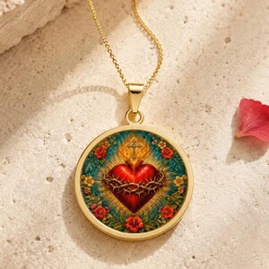 Puede incluir: Un collar de tono dorado con un colgante circular que presenta una imagen central de un corazón rojo rodeado por una corona de espinas, coronado con una cruz y enmarcado por un borde floral de hojas verdes y flores rojas sobre fondo azul.