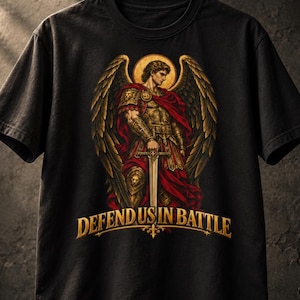 May include: A black short-sleeved t-shirt featuring a graphic of an angel with large wings, wearing gold armour and a red cape, holding a sword. Below the figure, the text DEFEND US IN BATTLE is printed in gold lettering with a decorative flourish.