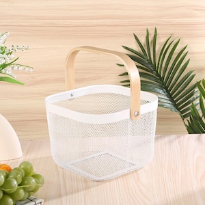 Pu&ograve; includere: Un cestino in filo metallico bianco con manico in legno. Il cestino ha una base quadrata e un bordo superiore curvo. Il manico &egrave; fissato al cestino con rivetti metallici. Lo sfondo comprende foglie verdi e una superficie in legno chiaro.