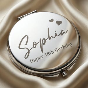 May include: A round, silver compact mirror with the name "Sophia" engraved in a cursive font, along with two heart symbols and the text "Happy 18th Birthday." The mirror is set against a satin background.