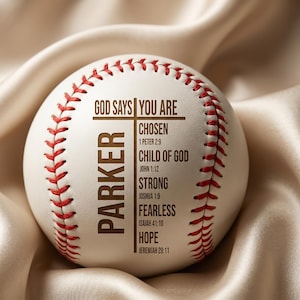 May include: A white baseball with red stitching and the text "GOD SAYS YOU ARE" and "PARKER". Additional text includes "CHOSEN", "CHILD OF GOD", "STRONG", "FEARLESS", and "HOPE" with associated Bible verses. The baseball is on a satin surface.