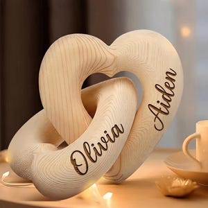 May include: A light-colored wooden sculpture consisting of two interlocking heart shapes. The surface of the wood displays a natural grain pattern. The name Olivia is engraved in a dark script font on one heart, and the name Aiden is engraved on the other.
